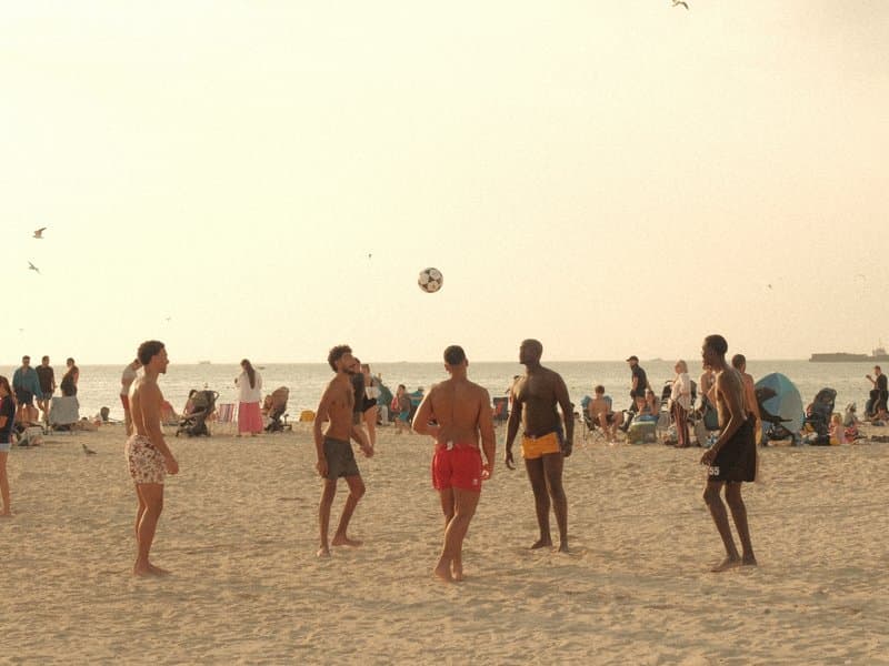 Group of people playing beach volleyball on a sandy beach with a hazy sky and distant beachgoers.