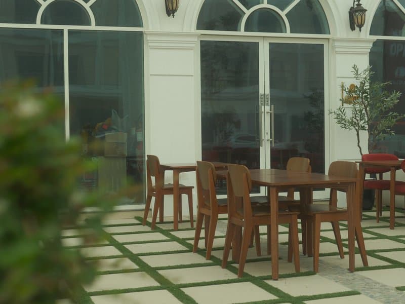 Outdoor dining area with wooden tables and chairs, surrounded by greenery and large windows.