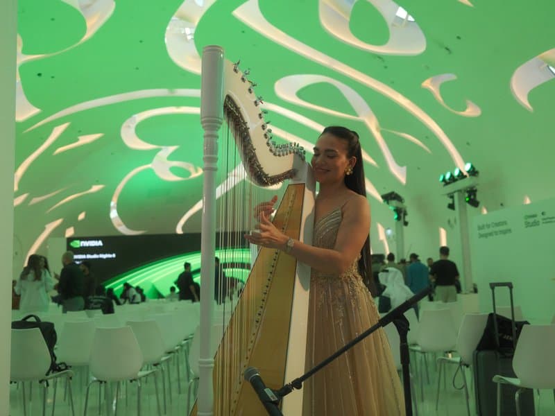 A woman in a golden dress plays a harp under a green-lit ceiling in a modern venue.