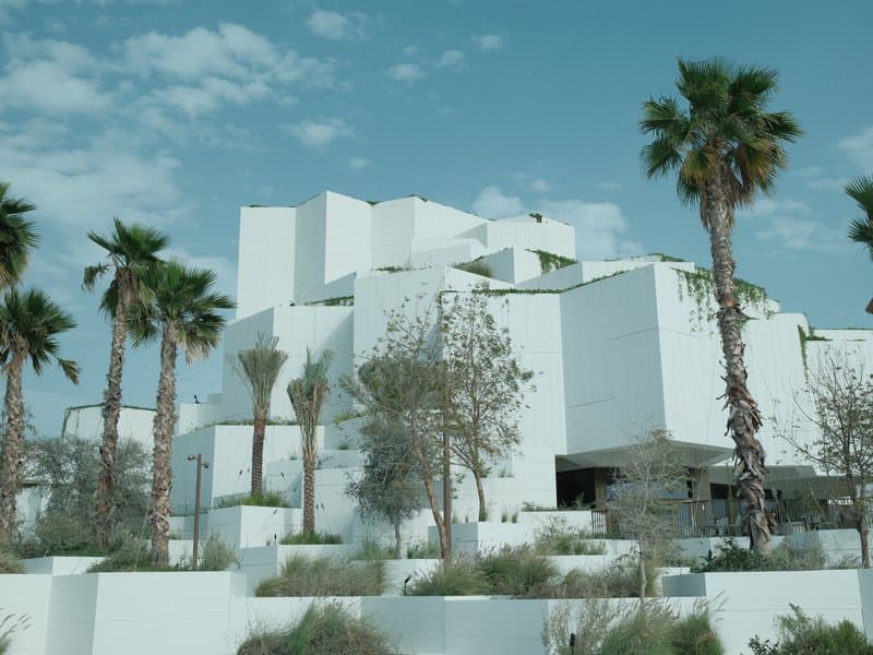 Modern white architectural building surrounded by palm trees and sparse vegetation under a blue sky.