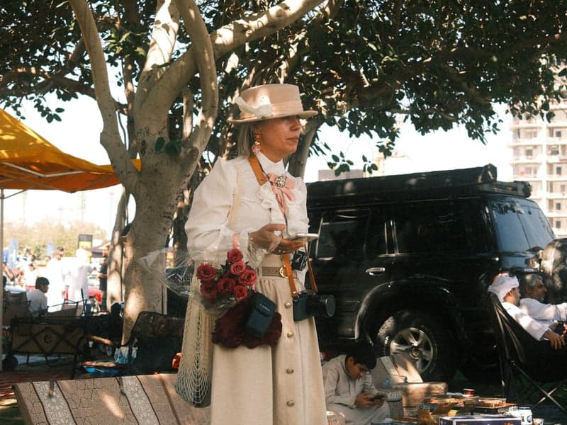 A woman in a white coat and hat stands outdoors, holding flowers, with a black vehicle in the background.