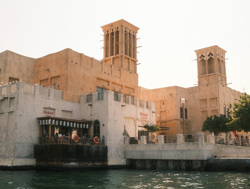 Historic building with wind towers by the water, people enjoying the view under warm sunlight.