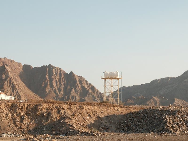 A water tower stands against a backdrop of rocky mountains under a clear sky.