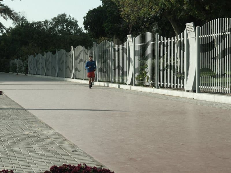 A person riding a scooter on a wide, empty pathway lined with a decorative fence and greenery.