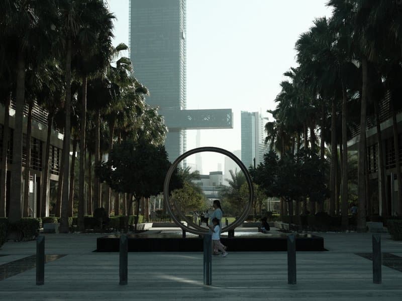A modern urban plaza with palm trees and a circular sculpture, surrounded by tall buildings.