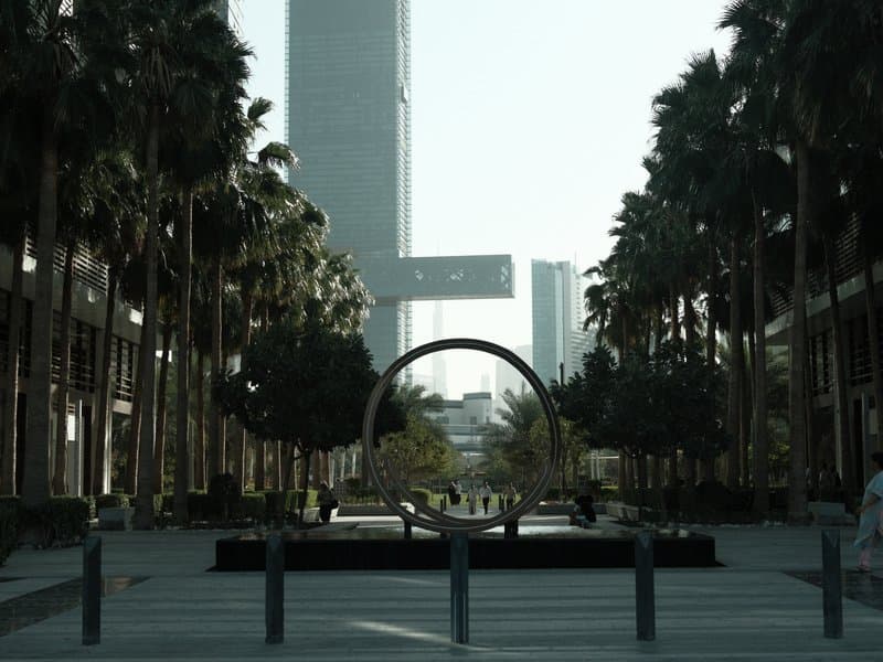 A modern urban plaza lined with palm trees and a circular sculpture, with skyscrapers in the background.