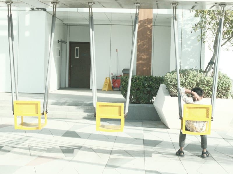 Two yellow swings in an urban setting with a person sitting on one swing, sunlight casting shadows.