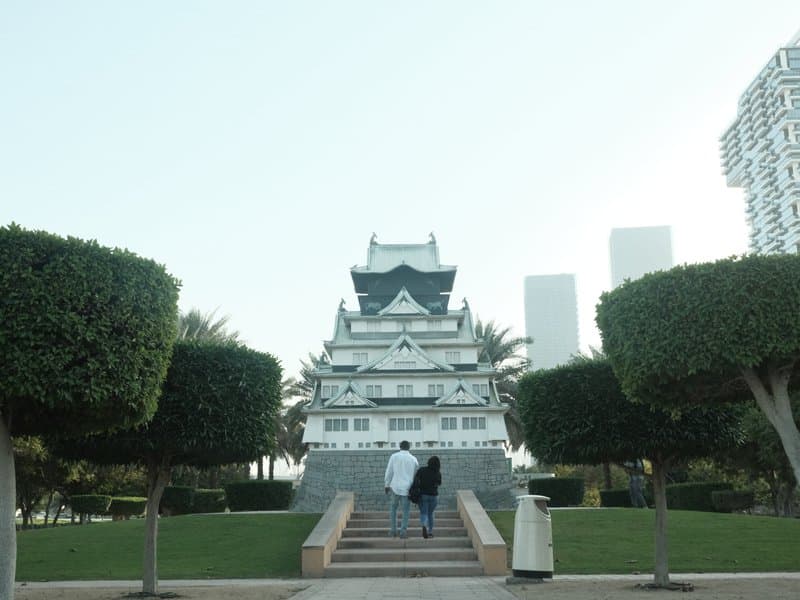 Two people walking towards a traditional-style building surrounded by neatly trimmed bushes and modern skyscrapers.