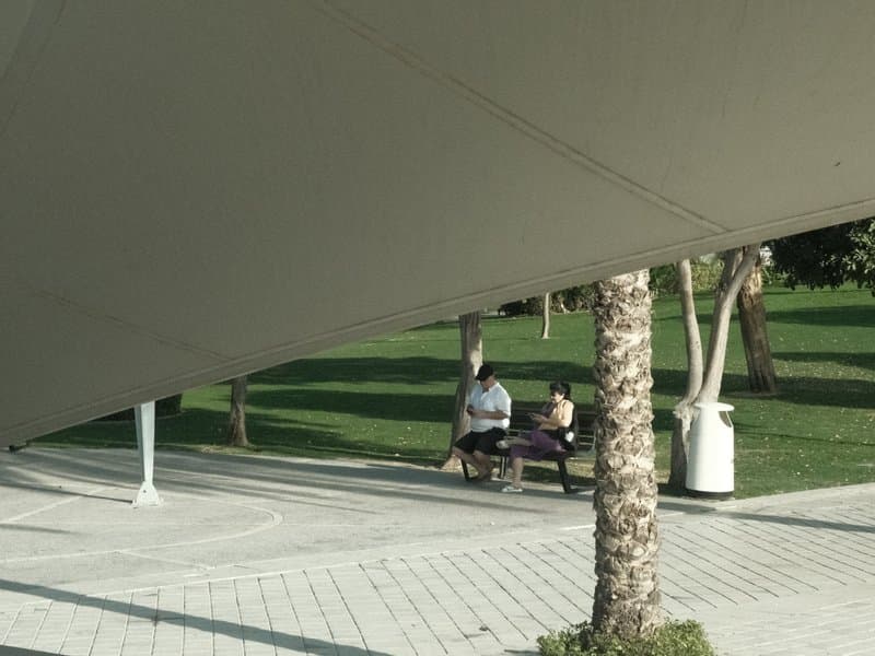Two people sitting on a bench in a park, partially obscured by a large canopy structure.