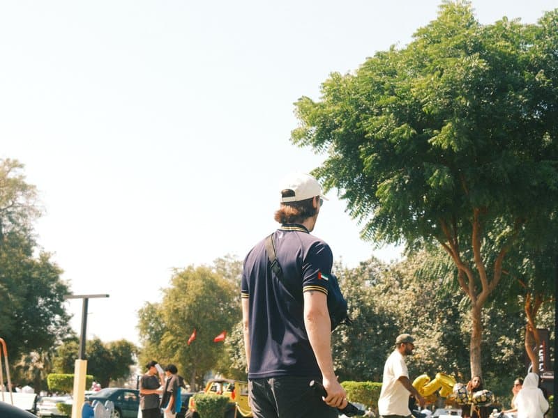 A person in a dark shirt stands with their back to the camera, surrounded by trees and people in a park.