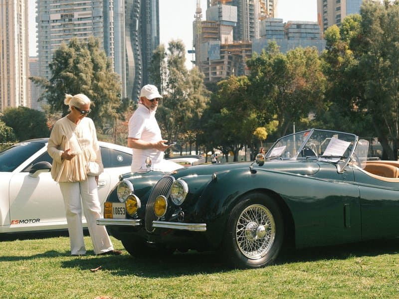 Two people admiring a classic green convertible car in a park with city buildings in the background.