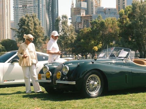 Two people admiring a classic green convertible car in a park with city buildings in the background.