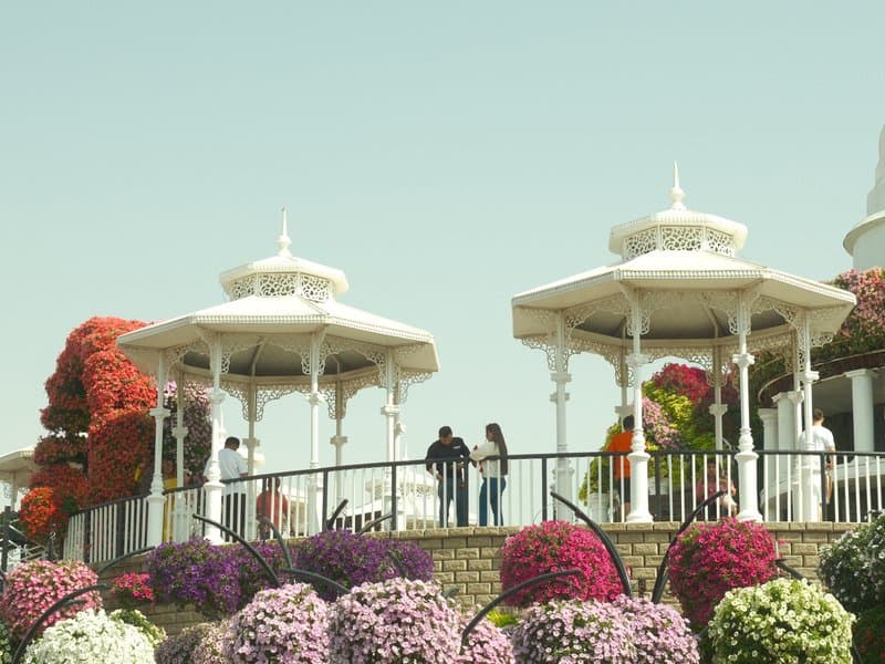 Two white gazebos surrounded by colorful flowers with people enjoying the scene.