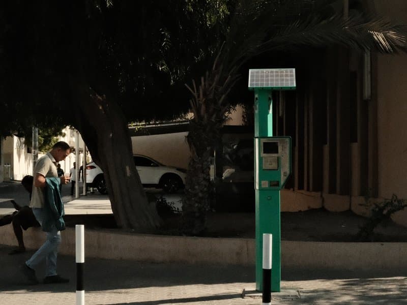 A man walking past a green parking meter under a tree in an urban setting.