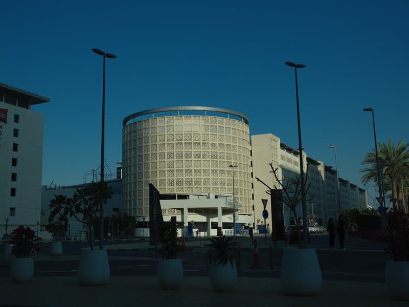 A modern building with a circular design under a clear blue sky, surrounded by palm trees and streetlights.