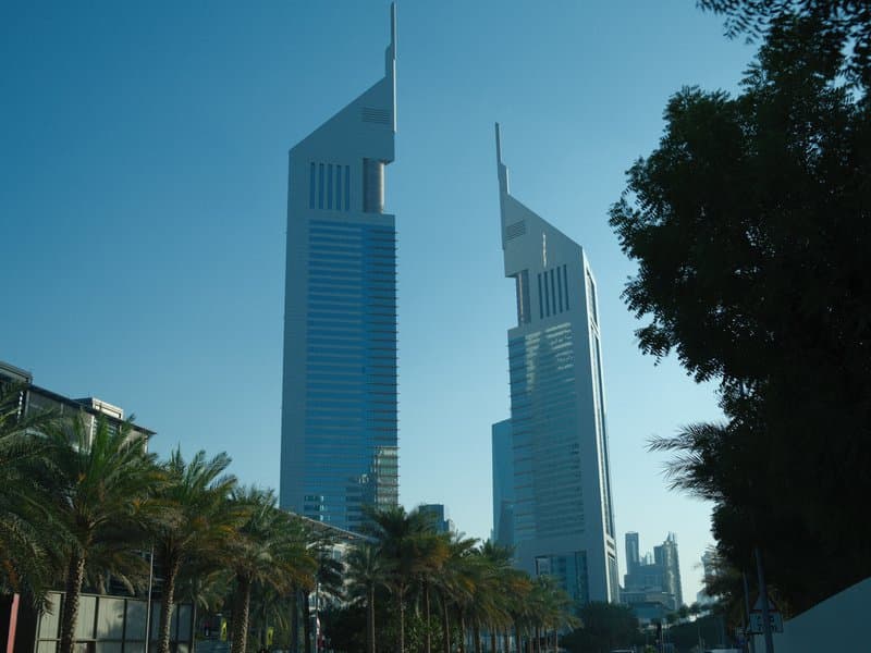 Two modern skyscrapers with pointed tops against a clear blue sky, surrounded by palm trees.