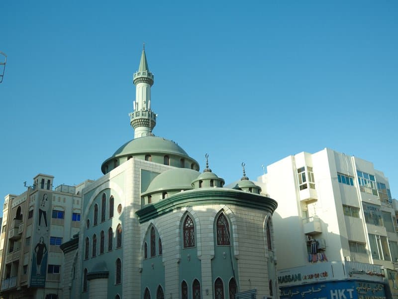 A green-domed mosque with a tall minaret against a clear blue sky, surrounded by modern buildings.