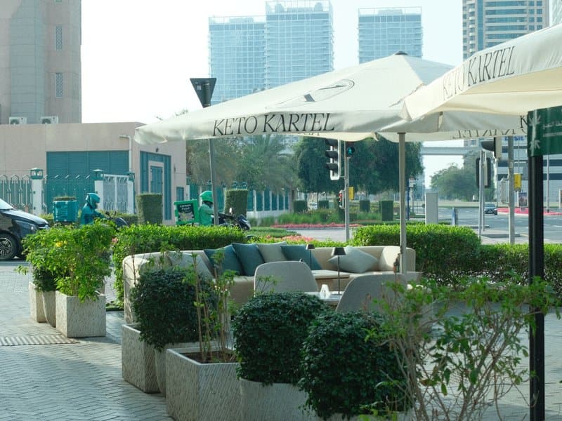 Outdoor café seating area with umbrellas and greenery, urban setting in the background.