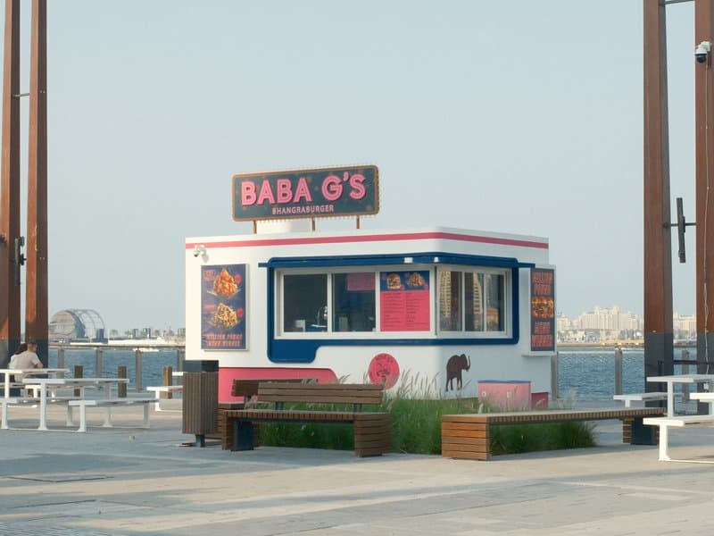A colorful food stall named Baba G's by the waterfront with a clear sky and distant city skyline.