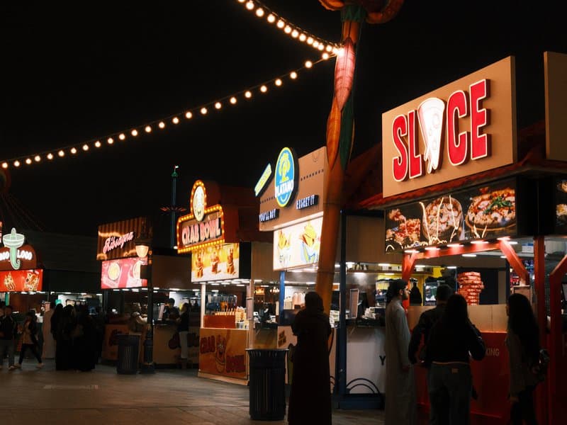 Nighttime scene of food stalls with bright signage and string lights in an outdoor setting.