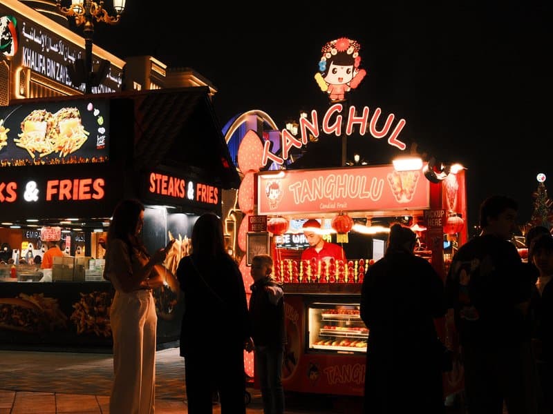 A vibrant food stall at night with people gathered around, illuminated by colorful lights.