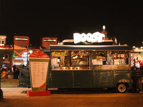 A food truck named 'Boost' illuminated at night with a menu board beside it, set in a lively outdoor area.