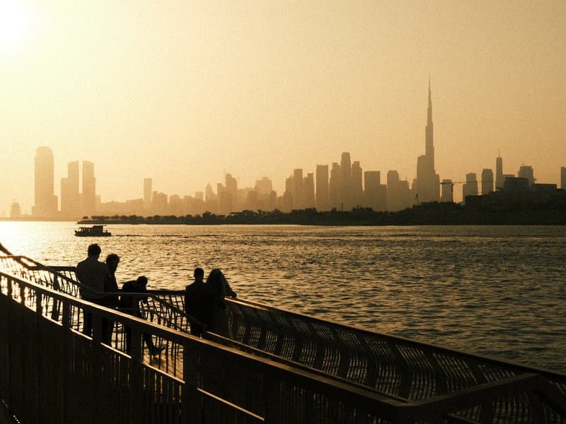 Silhouette of a city skyline at sunset with people on a pier overlooking the water.