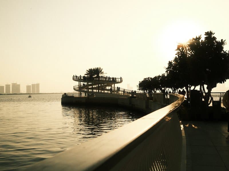 A serene waterfront scene with a winding path, trees, and a distant city skyline at sunset.