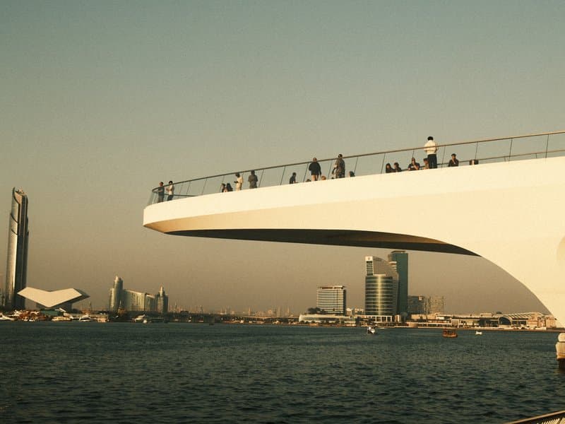 A modern bridge with people walking on it, overlooking a city skyline at sunset.