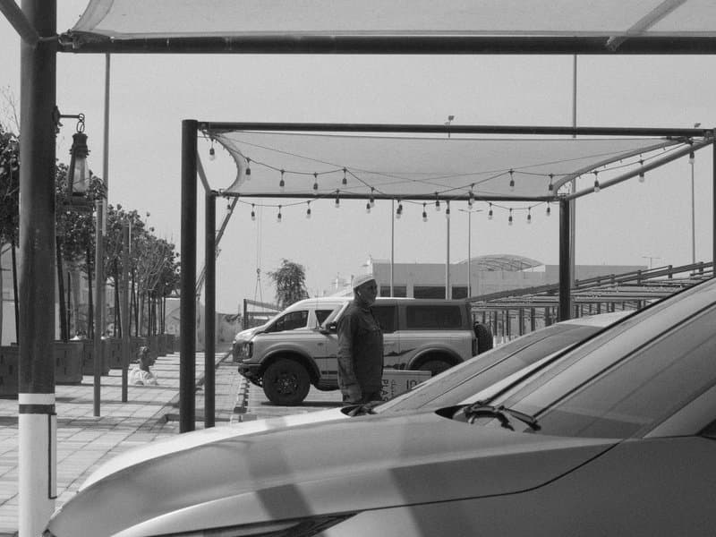 A man stands in a parking area with vehicles and a canopy overhead, in a monochrome setting.