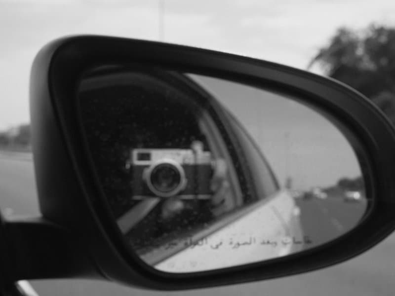 A black and white image of a side mirror reflecting a camera and a road.