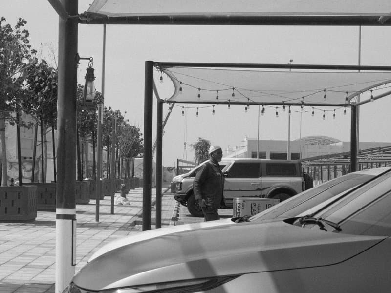 A black and white street scene featuring a man standing near parked cars under a canopy.