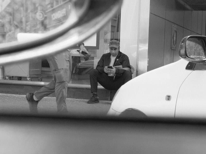 A man sitting on the sidewalk, looking at his phone, with a blurred figure walking past in the foreground.