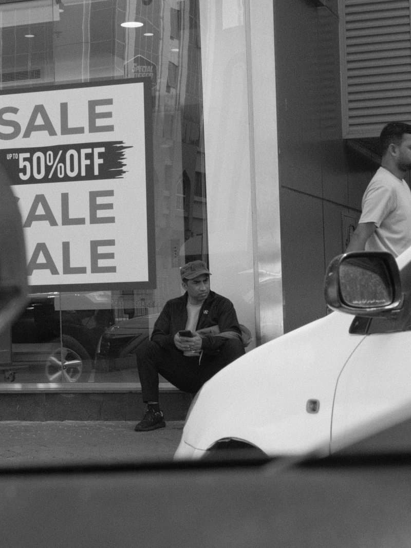 A man sitting on the sidewalk near a store with a sale sign, another person walking by.