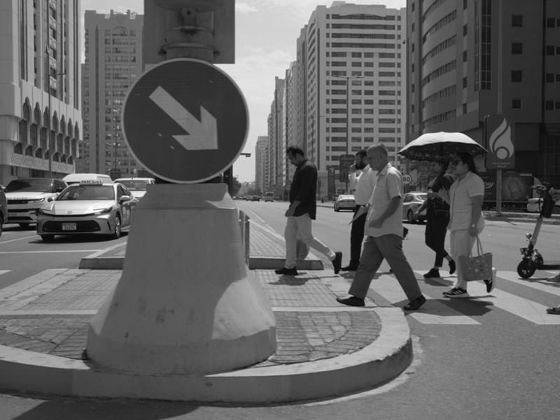 People crossing a street in an urban setting with a traffic sign and tall buildings in the background.