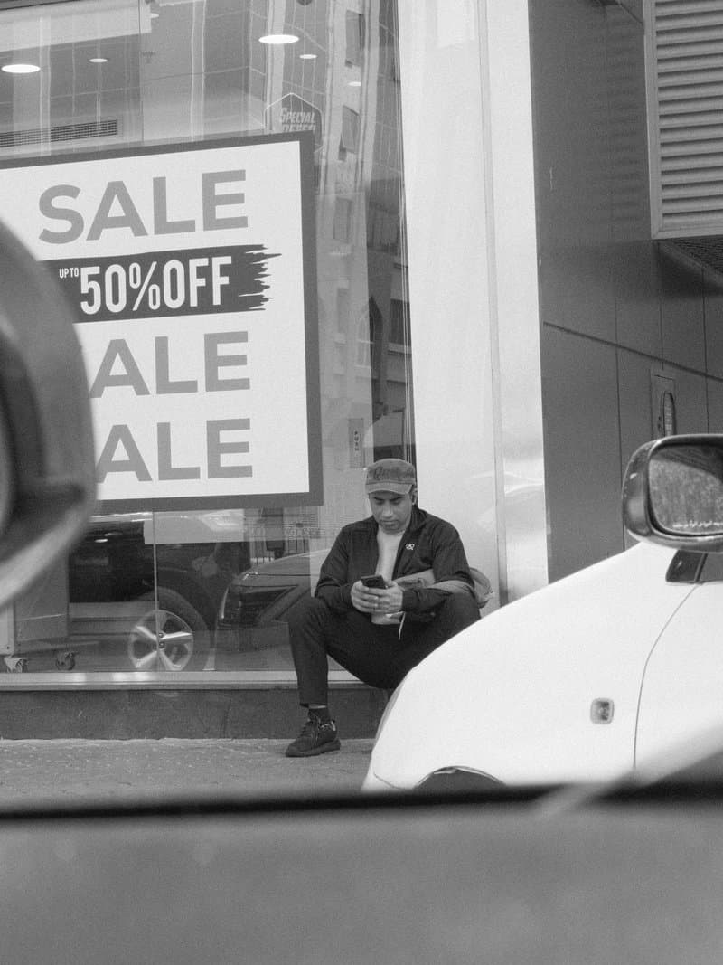 A man sitting outside a store with a large sale sign, looking at his phone.