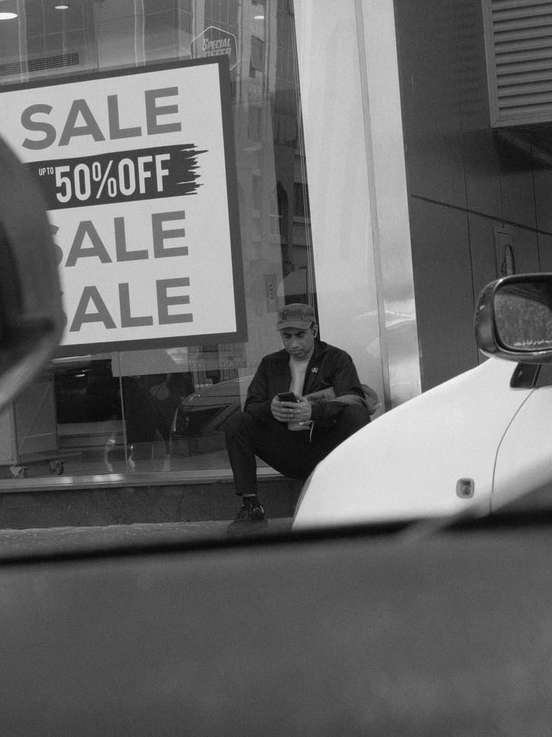 A man sitting on the sidewalk near a store with a sale sign, looking at his phone.