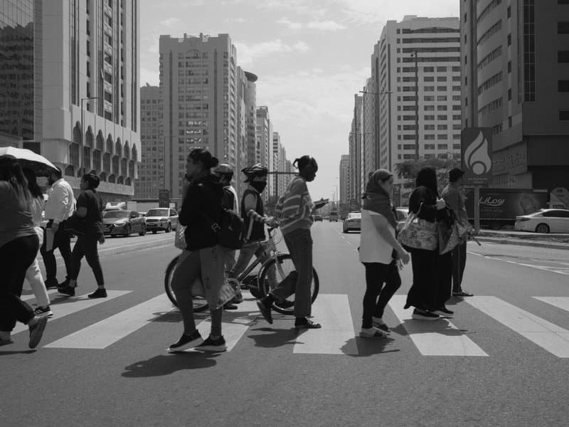 Black and white image of pedestrians crossing a street with tall buildings in the background.