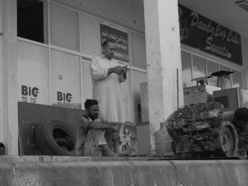 Two men outside a shop, one standing and one sitting near a vehicle engine and tires.
