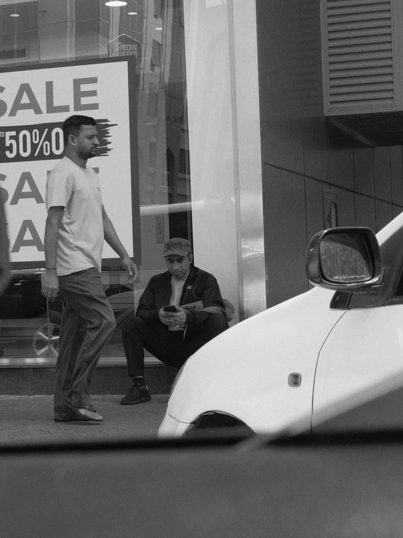A street scene featuring two men, one standing and one sitting, with a sale sign in the background.