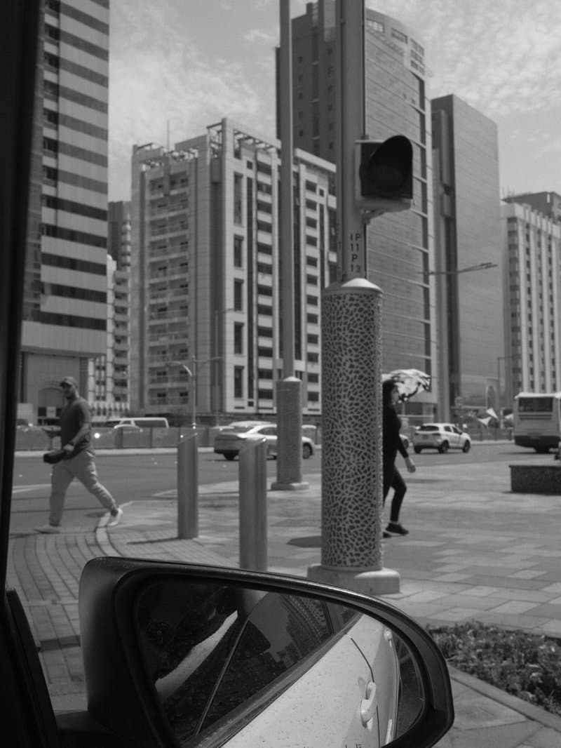 Black and white urban scene showing pedestrians crossing near a traffic signal with modern buildings in the background.