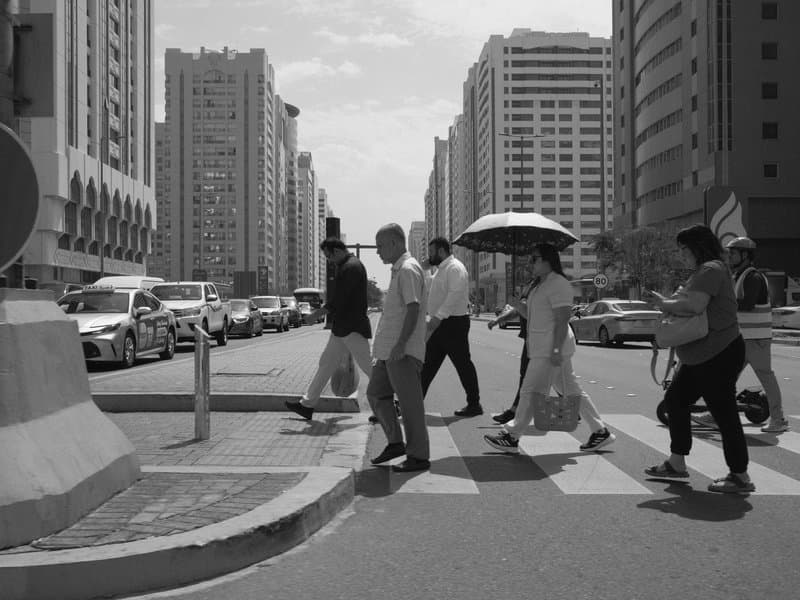 Black and white street scene of pedestrians crossing a busy urban intersection with tall buildings in the background.