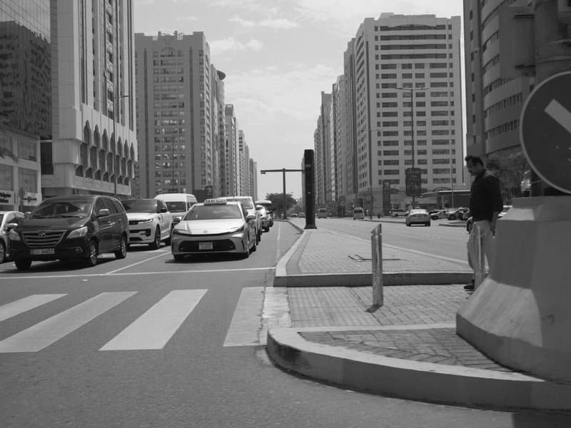 A busy urban street scene with cars, tall buildings, and a pedestrian waiting at a crosswalk.