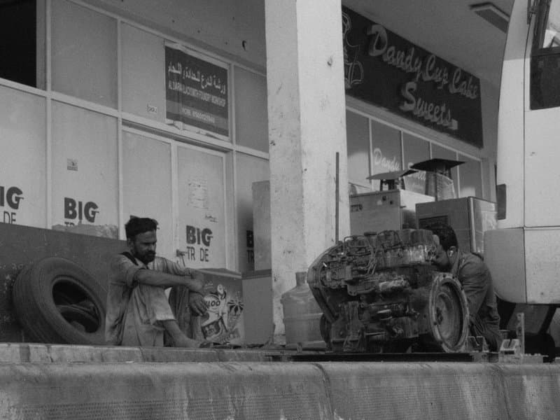 A man sitting beside a vehicle engine in an urban setting with storefronts in the background.