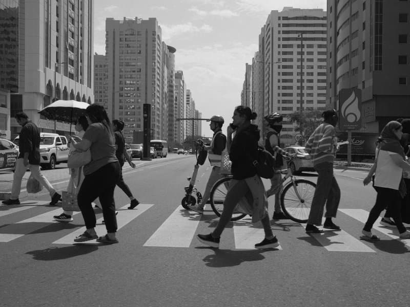 Black and white photo of pedestrians crossing a busy city street with tall buildings in the background.