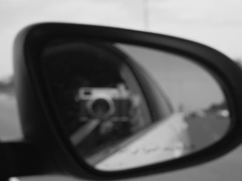 A black and white image of a side mirror reflecting a camera and a blurred road.