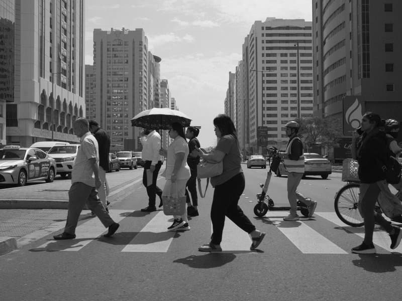 Black and white street scene of pedestrians crossing a busy urban intersection with tall buildings.