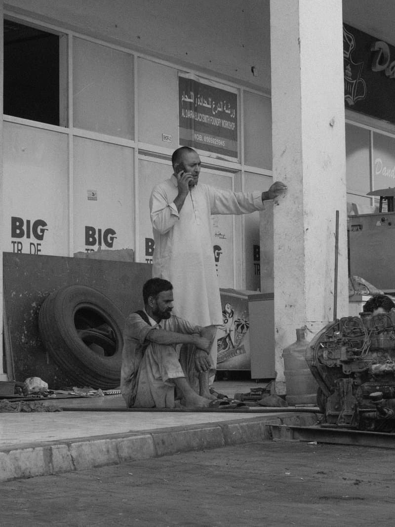 Two men in casual clothing, one standing and one sitting, near a storefront with tires and signage.