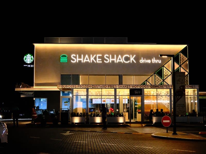 Night view of a Shake Shack restaurant with bright signage and a Starbucks nearby.