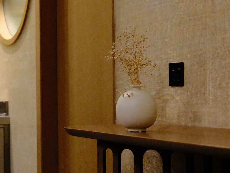 A minimalist interior scene featuring a white vase with dried flowers on a wooden shelf against a textured wall.